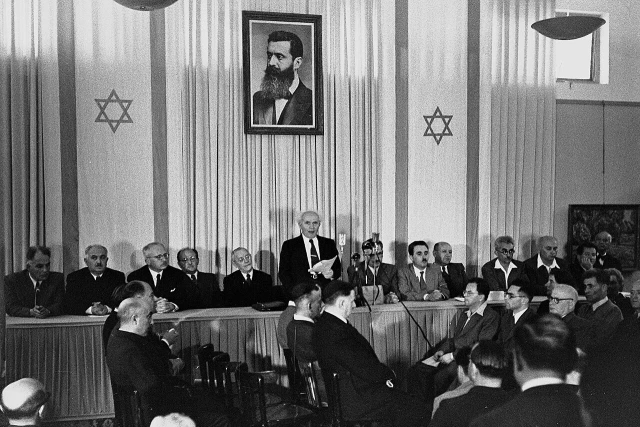 David Ben-Gurion publicly declares the establishment of the State of Israel on May 14, 1948, in Tel Aviv, beneath a portrait of Theodor Herzl, the founder of modern political Zionism, at the former Tel Aviv Museum of Art on Rothschild Street. (Photo: Wikimedia Commons)