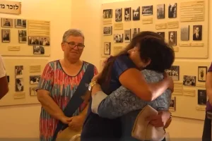 Miriam, the owner of the Beit Shalom guesthouse, embraces one of the German volunteers in Metula, beside a memorial display honoring her relatives murdered in the Holocaust. (Screenshot: Kan 11)